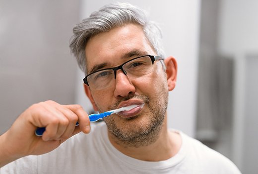 A middle-aged man wearing glasses and brushing his teeth while standing in front of the mirror
